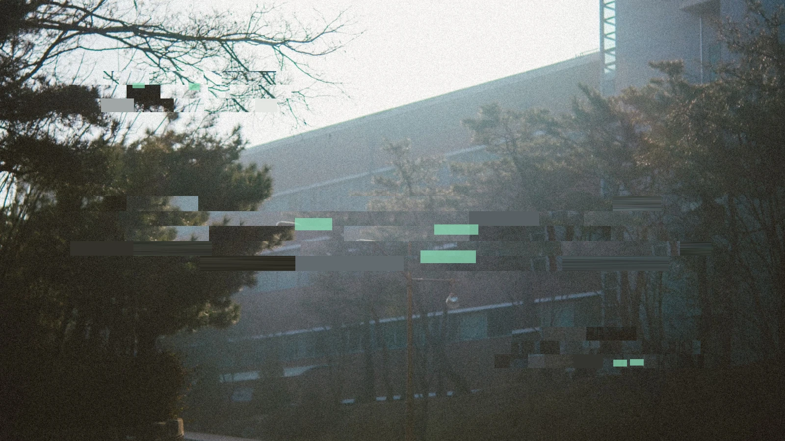A large building partly concealed by trees and haze, interrupted by broad digital fragments and small coloured blocks.